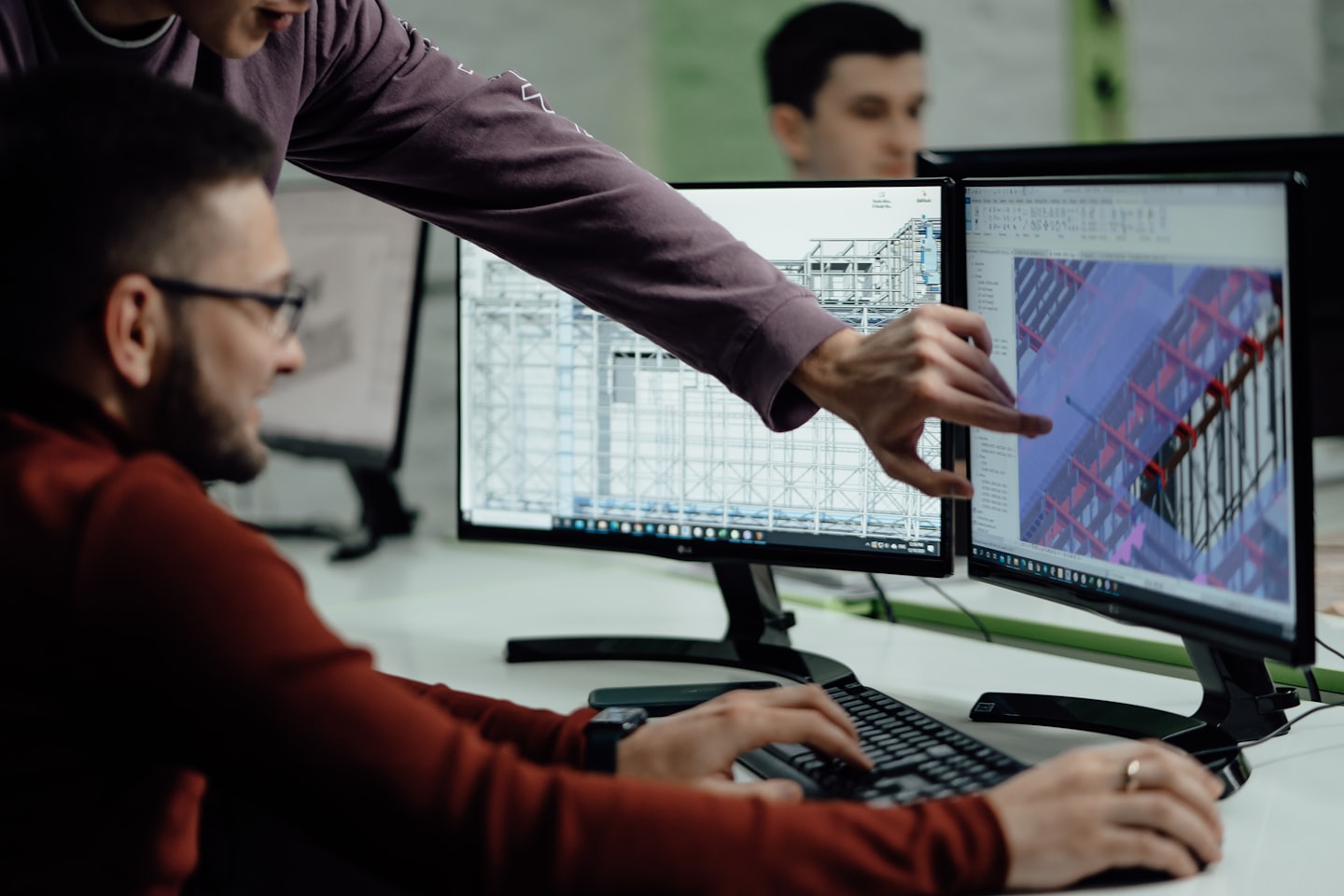 Mann zeigt auf 3D-Design auf zwei Monitoren, während ein Kollege tippt.